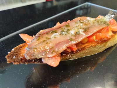 de mesa en mesa-Tostada de pan con tomate macerado aceite y hierbas y encima una anchoa del cantábrico